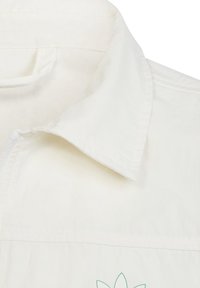 Bílá, strukturovaná látková košile s límečkem a předním zipem; má zelený květinový vzor blízko spodního lemu. Čistý a minimalistický design.