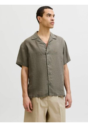 Homme portant une chemise khaki à manches courtes boutonnée et un pantalon beige, debout devant un fond clair uni, regardant sur le côté.