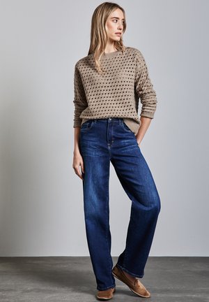 Beige knitted sweater with open dot pattern, paired with dark blue wide-leg jeans and light brown slip-on shoes. Subtle textures throughout.