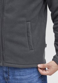 Fleece jacket in dark gray, featuring a full zipper and side pockets. The fabric has a soft, textured appearance. Reinforced hem with brand tag.
