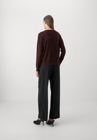 Femme aux longs cheveux bruns portant un pull bordeaux foncé, un pantalon noir à jambes larges et des mules noires à talons, debout devant un fond clair uni.
