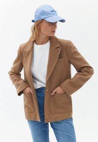 Hnědá blazer z vlněné směsi s klopovým límcem, dvěma předními kapsami a zapínáním na jeden knoflík, kombinovaná s bílým tričkem a modrými džíny.