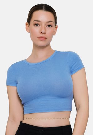 Młoda kobieta ubrana w dopasowany jasnoniebieski krótki top i czarne spodnie, stojąca na jednolitym białym tle.