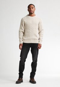 Beige kabelstickad tröja med rund halsringning, strukturerat mönster, figurnära stil. Matchad med mörka jeans och bruna snörskor.