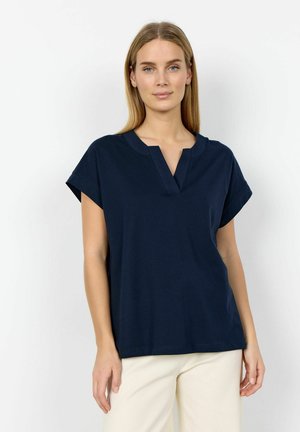 Frau mit glattem blondem Haar, die ein marineblaues kurzärmeliges Oberteil mit einem V-Ausschnitt mit Ausschnitt und hellfarbene Hosen trägt und vor einem weißen Hintergrund steht.