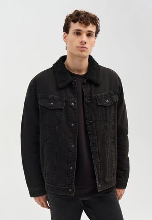 Giovane uomo che indossa una giacca di jeans nera con colletto sherpa nero e pantaloni neri abbinati su uno sfondo chiaro e semplice.
