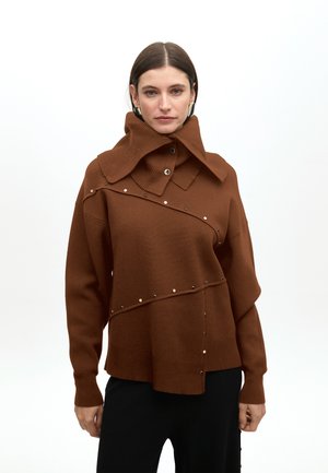 Suéter marrón de punto oversize con un cuello alto, que presenta una línea diagonal de tachuelas de metal decorativas y un diseño de corte holgado.