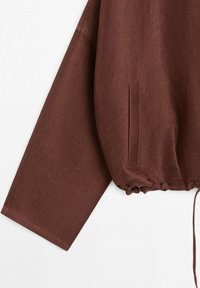 Veste marron à manches longues avec poche latérale fendue et ourlet à cordon de serrage sur fond blanc.