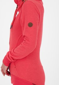 Coral hoodie with a zippered front, ribbed cuffs, and hem. Features a small brown logo patch on the left sleeve. Soft texture and fitted design.