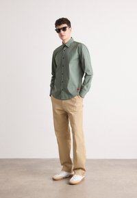 Cămașă verde cu mâneci lungi și închidere cu nasturi, combinată cu pantaloni bej și adidași albi. Cămașa are un detaliu cu etichetă roșie.