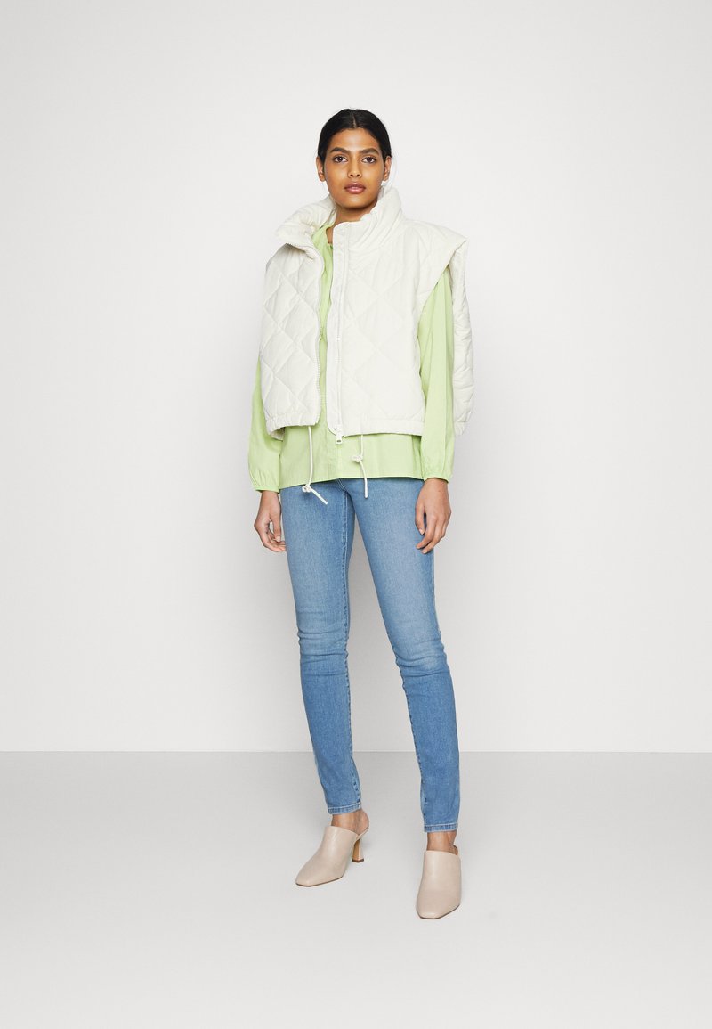 Kurtka z pikowanej ecru tkaniny z wysokim kołnierzem, noszona na jasnobrązowej bluzce. Modelka zestawia ją z niebieskimi obcisłymi dżinsami oraz beżowymi klapkami na obcasie.