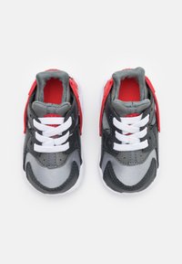 Par de pequenos sapatos desportivos com malha cinza, sobreposições pretas, tiras ajustáveis vermelhas, atacadores brancos e forro interior acolchoado vermelho.