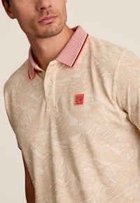 Crème polo shirt met subtiel bladerenpatroon, contrasterende gestreepte kraag in rood en wit, knoopsluiting en een gestructureerde stofafwerking.