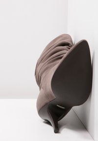 Scarpa grigia in suede con tacco alto, punta affilata e texture arricciata, dotata di un tacco scuro a punta e suola liscia.