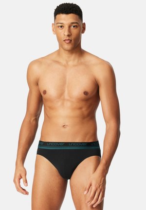 Svarta bomullsbriefs med en bred elastisk midjeresår med texten "uncover" i teal. Designen är slät och kroppsnära.