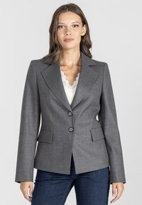 Kobieta w dopasowanej szarej marynarce, nosząca białą koronkową bluzkę i ciemnoniebieskie dżinsy, stojąca na tle jasnego, jednolitego tła.