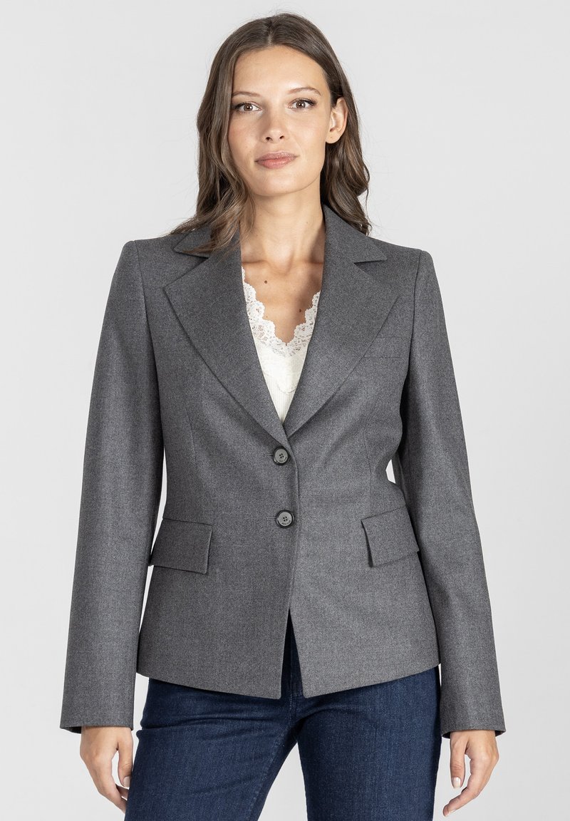 Kobieta w dopasowanej szarej marynarce, nosząca białą koronkową bluzkę i ciemnoniebieskie dżinsy, stojąca na tle jasnego, jednolitego tła.