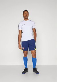 Mlad moški stoji oblečen v belo športno majico Nike, modre kratke hlače Atletico Madrid, modre nogavice in črne nogometne kopalke.