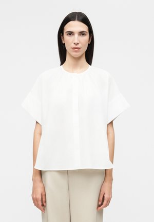 Femme aux cheveux raides et foncés portant un chemisier blanc ample à manches courtes et un pantalon beige sur un fond clair uni.