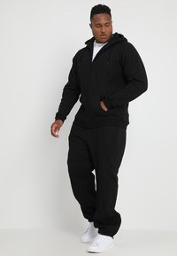 Black zip-up hoodie with front pockets and adjustable drawstring. Paired with matching black sweatpants. White sneakers complete the look.