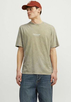 T-shirt en coton vert olive clair avec une coupe décontractée, des manches courtes et une impression sur le devant indiquant "Originals Studio". Associé à un jean en denim bleu.