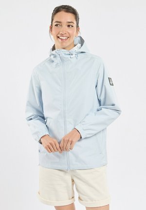 Femme souriante portant une veste à capuche bleu clair et un short crème, debout devant un fond blanc uni.