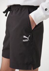 Personne portant un short noir Puma avec une taille élastique et la main dans la poche, associé à un t-shirt blanc à manches longues affichant un logo sur la manche.