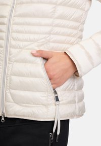 Lichte pufferjack in crème met een verticale doorgestikte ontwerp, voorzien van een zilveren rits en een zijzak met een trekkoordverstelling.