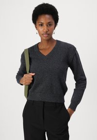 Maglione in misto lana grigio con scollo a V, polsini e orlo a costine. Indossato con pantaloni neri su misura, caratterizzati da una tracolla verde oliva.
