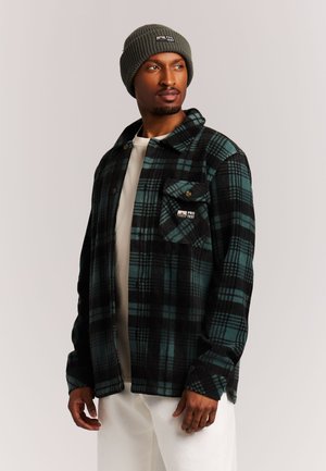 Hombre con gorro de punto gris y camisa de cuadros verde y negra abotonada sobre pantalones blancos, mirando hacia un lado.