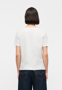 Γυναίκα με κοντά σκούρα μαλλιά, φοράει απλό λευκό κοντομάνικο πουκάμισο και σκούρο μπλε τζιν, στραμμένη προς τα πίσω με φόντο λευκό.