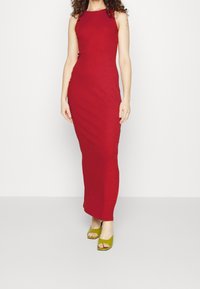Rotes ärmelloses Maxikleid aus glattem Stoff mit einer taillierten Silhouette. Kombiniert mit grünen offenen Schuhen mit Absatz, die ein schlichtes und modernes Design präsentieren.