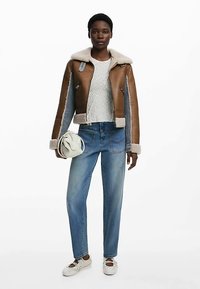 Giacca di pelle marrone con fodera in shearling, maglione bianco lavorato a maglia, jeans azzurri e scarpe da ginnastica bianche. La modella tiene una borsa crema.