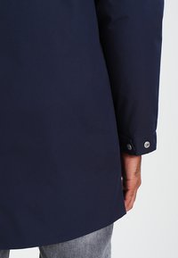 Cappotto impermeabile blu navy con taglio dritto, caratterizzato da chiusure a scatto sui polsini e un tessuto dalla texture liscia.