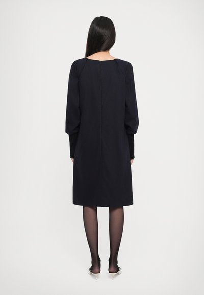 Woman with long dark hair wearing a black knee-length dress, sheer black tights, and white pointed heels, standing facing a plain white background.
