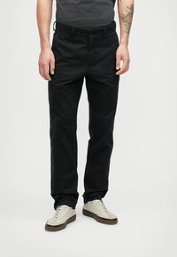 Άνδρας που φοράει στενά μαύρα παντελόνια και ανοιχτόχρωμα αθλητικά παπούτσια, στέκεται μπροστά σε καθαρό λευκό φόντο.