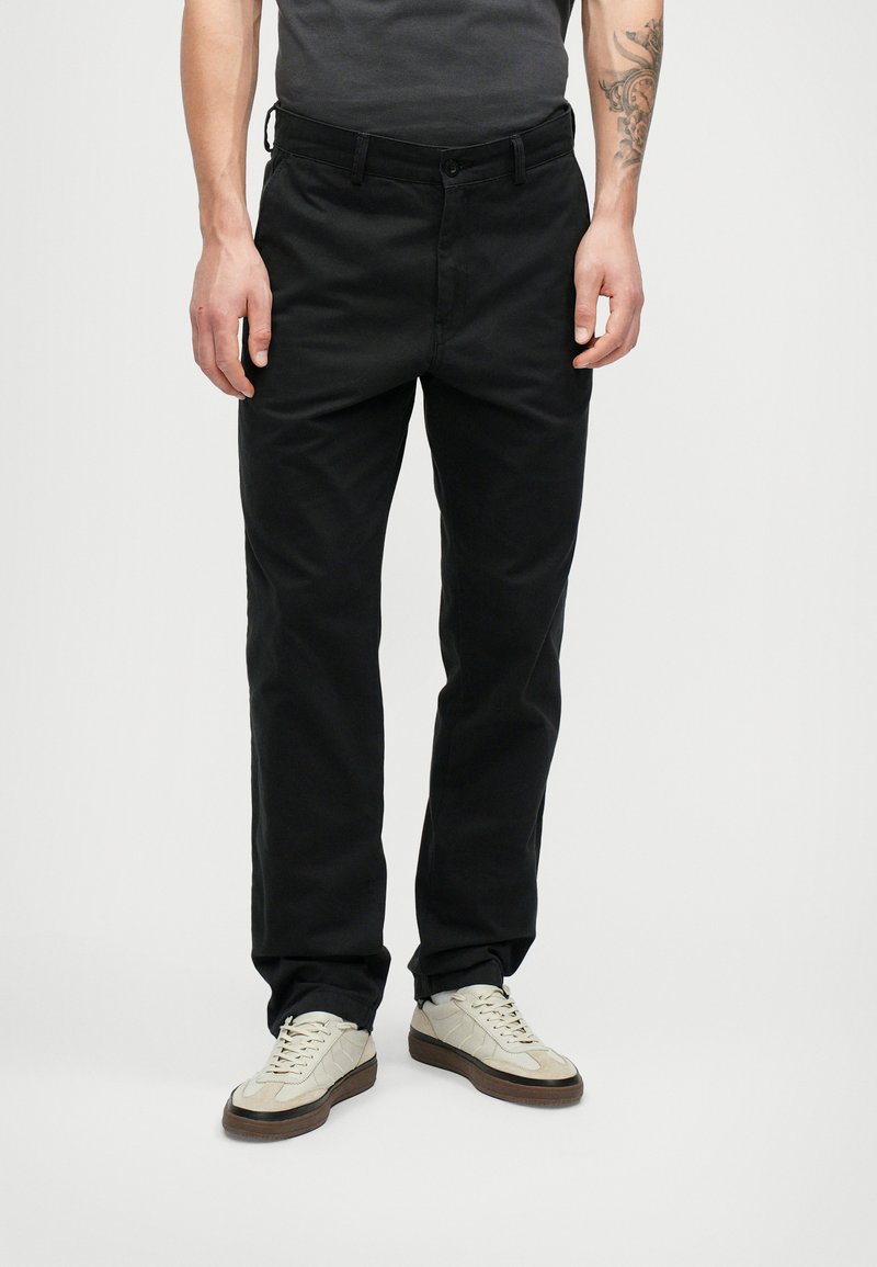 Άνδρας που φοράει στενά μαύρα παντελόνια και ανοιχτόχρωμα αθλητικά παπούτσια, στέκεται μπροστά σε καθαρό λευκό φόντο.