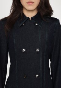 Mørkeblå denimjakke med dobbeltradet design, grønn søm, store svarte knapper og spiss krage.