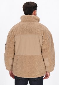 Mand, iført beige sherpa fleecejakke med nylonpanel på ryggen og ærmerne, står med ryggen mod kameraet mod en hvid baggrund.