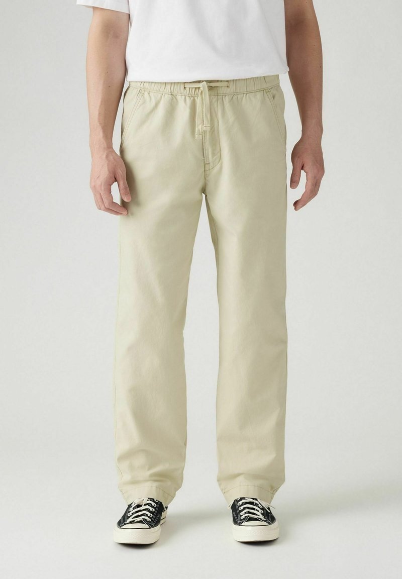 Pantalones de algodón beige claro con cintura elástica, piernas rectas y bolsillos laterales, combinados con zapatillas negras y blancas.
