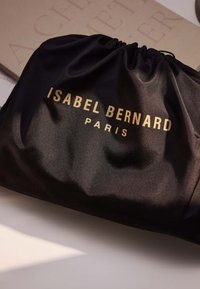 Černý stahovací pytel vyrobený z hladké tkaniny, na němž je vyražený zlatý nápis: "ISABEL BERNARD PARIS." Jednoduchý, elegantní design s minimální texturou.
