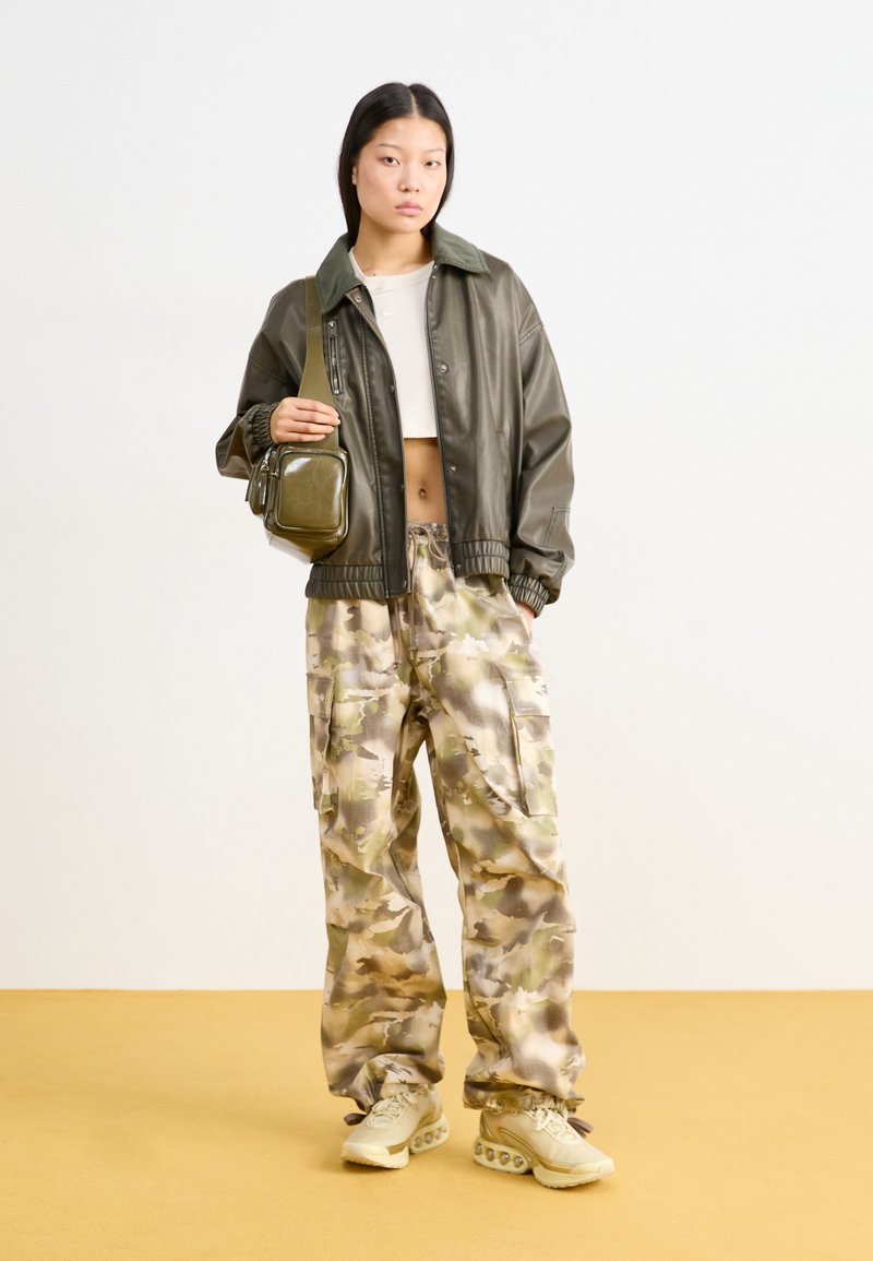 Olivgrüne Bomberjacke, verkürztes weißes Top, camo-musterte, weite Hose und beige Sneakers mit sichtbarem Polster und strukturierten Sohlen.