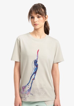 T-shirt en coton beige présentant un graphique coloré d'une figure avec des accents bleus et roses, tendant la main, avec un léger effet de éclaboussures en bas.