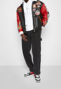 Man med svart och röd läderjacka med lappar, vit huvtröja, svarta jeans och svarta, vita och röda sneakers som står mot en neutral bakgrund.