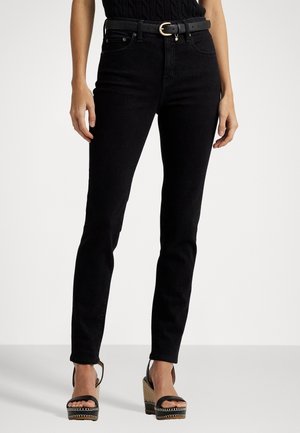 Femme portant un jean skinny taille haute noir avec une ceinture noire et des sandales compensées noires, debout devant un fond uni.
