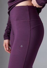 Leggings de entrenamiento en un profundo color púrpura con una textura suave, que presentan costuras planas y un pequeño logo cerca de la cintura, ofreciendo un ajuste aerodinámico.