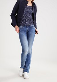 Dunkelblaue Jacke über einem gemusterten marineblauen Tanktop, kombiniert mit hellblauen Jeans und weißen Sneakers; lässiger Stil mit einer figurbetonten Silhouette.