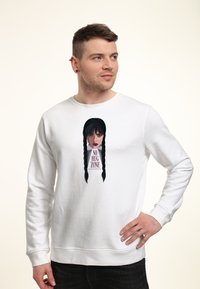Uomo con capelli corti castani e orecchini indossa un sudario bianco con l'immagine di una donna dai capelli scuri con trecce e il testo "NO HUG ZONE."