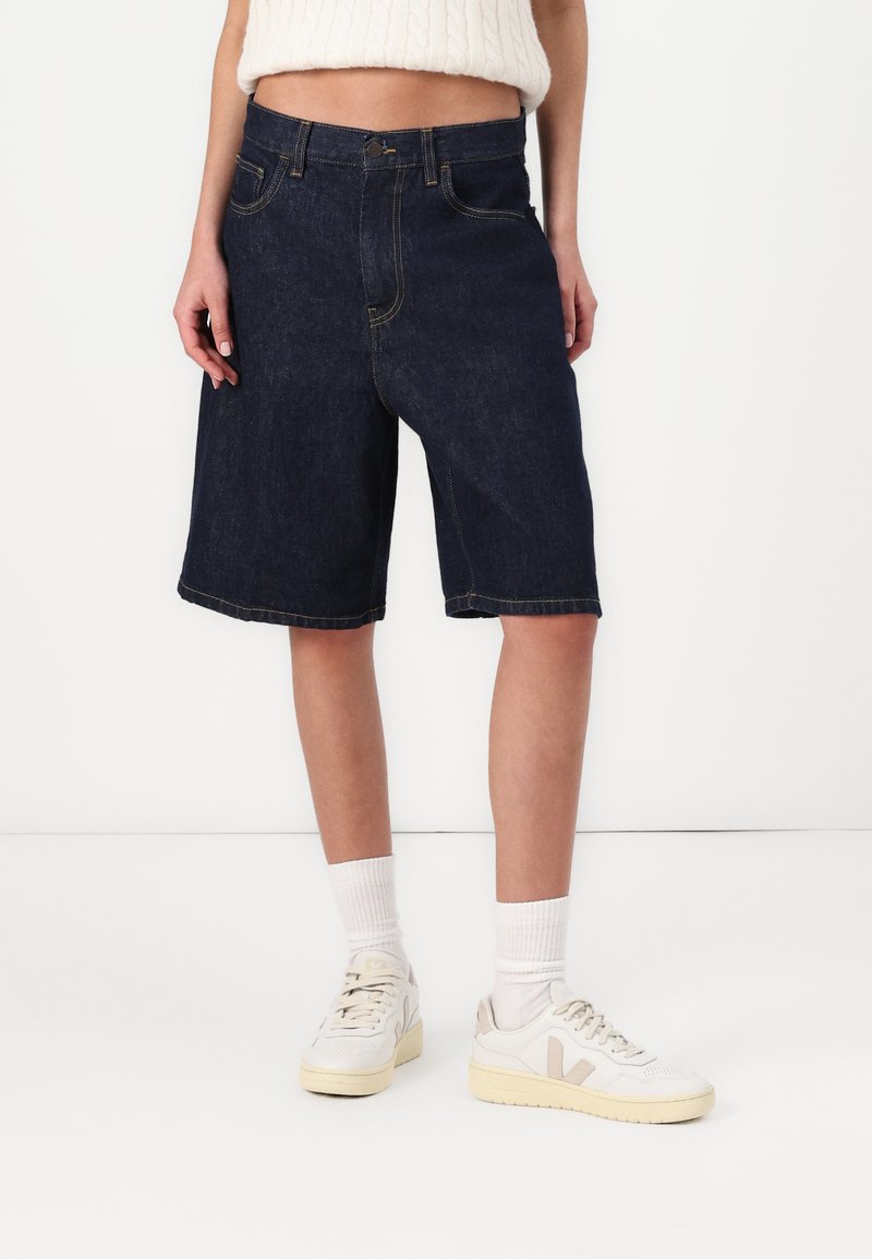 Shorts en denim bleu marine avec une coupe décontractée, dotés de cinq poches, passants de ceinture et coutures jaunes contrastantes ; associés à des baskets blanches.