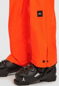 Oranje skibroek met zwarte hardware en logo, gedragen over zwarte skischuilen met verstelbare gespen en textuur oppervlaktes.